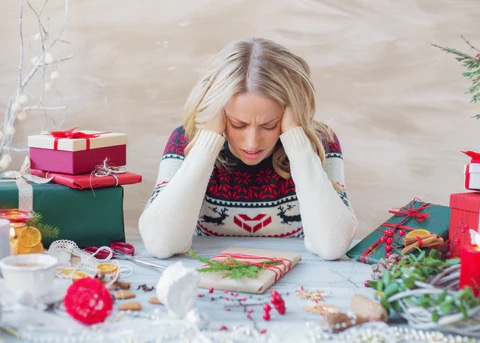 Overwhelmed woman sitting at a table with wrapped Christmas gifts and holiday clutter