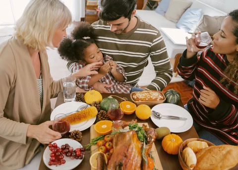 a standing man holding a child at the Thanksgiving table talking to two women, also standing